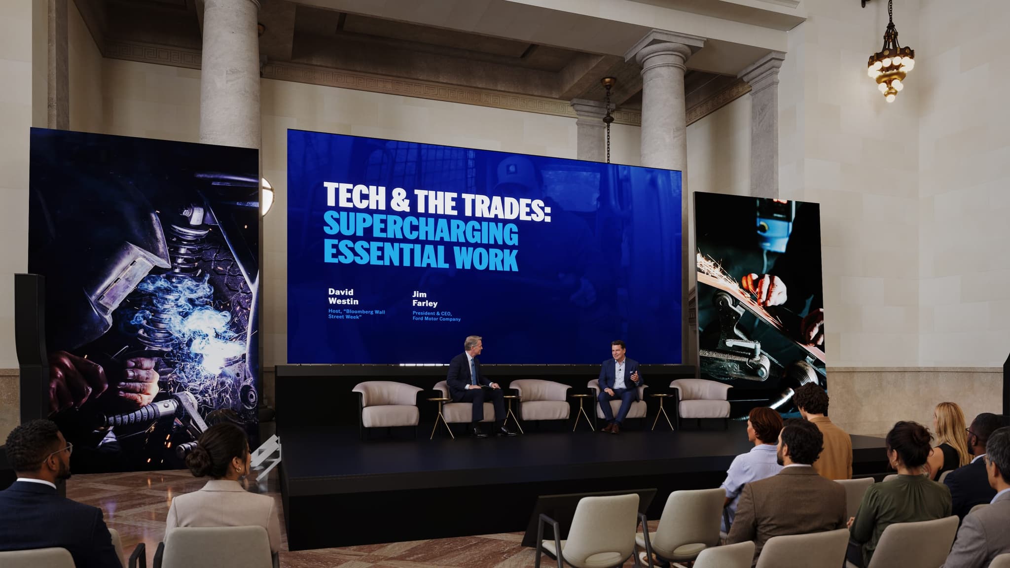 Closer render of the Grand Hall stage at Michigan Central Station, showing a panel discussion on a low stage in front of a large screen, with tall screens on either side and the stone columns behind.